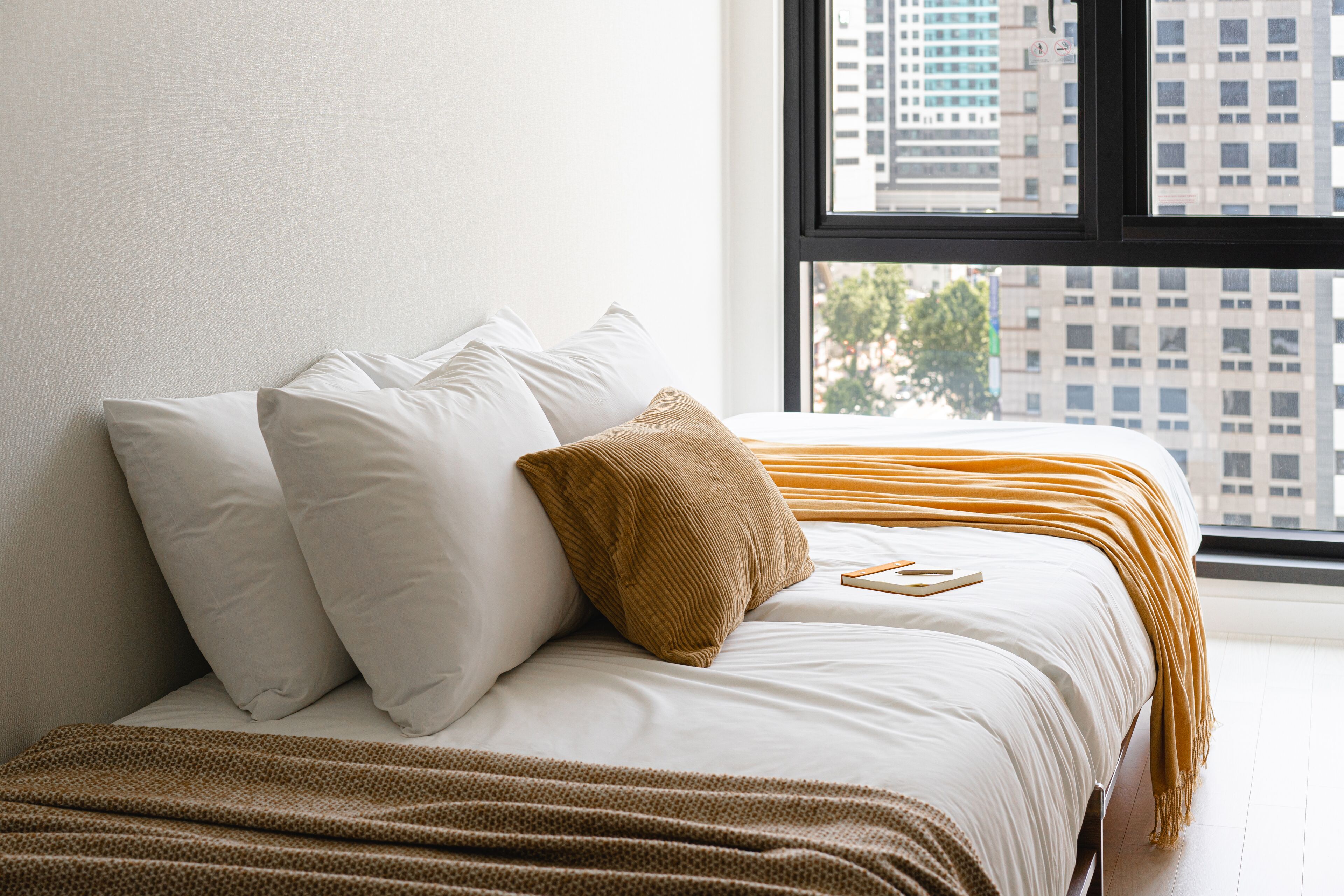 A neatly made bed with white and brown bedding, a window with a city view, and a plain wall.