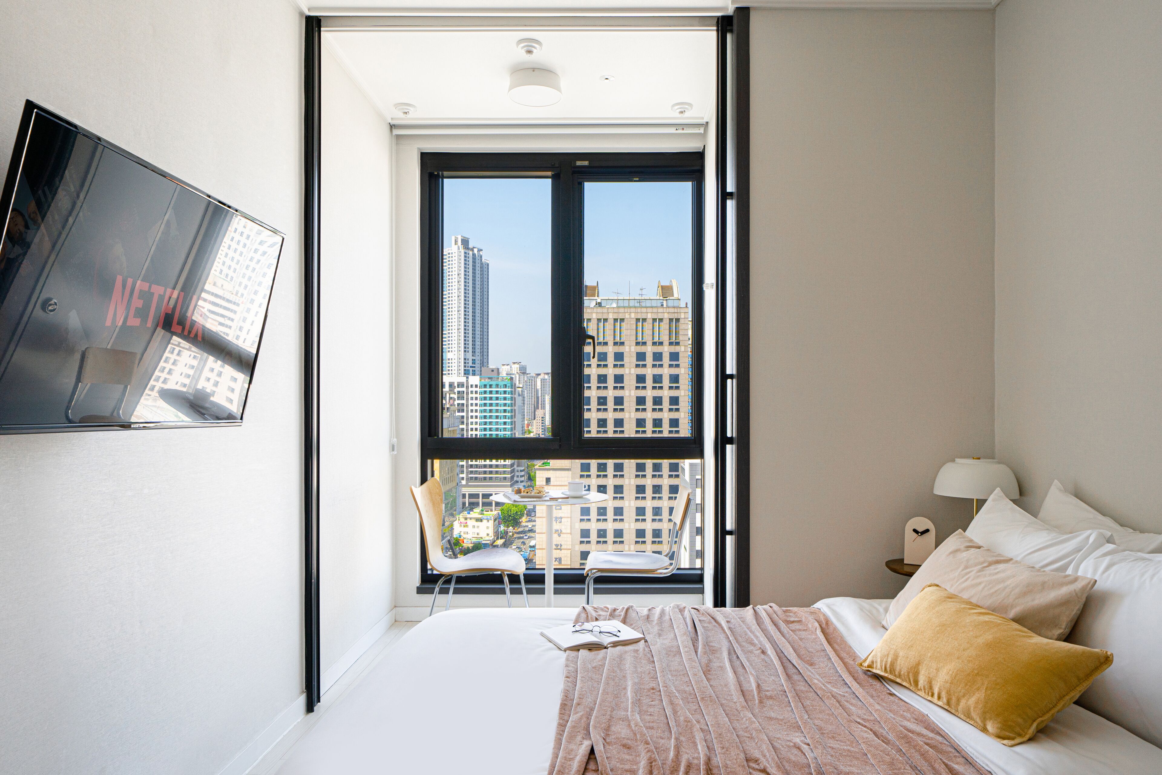 Une chambre d’hôtel moderne équipée d’un lit, d’un téléviseur fixé au mur, d’un balcon offrant une vue sur la ville et d’une petite table avec des chaises.