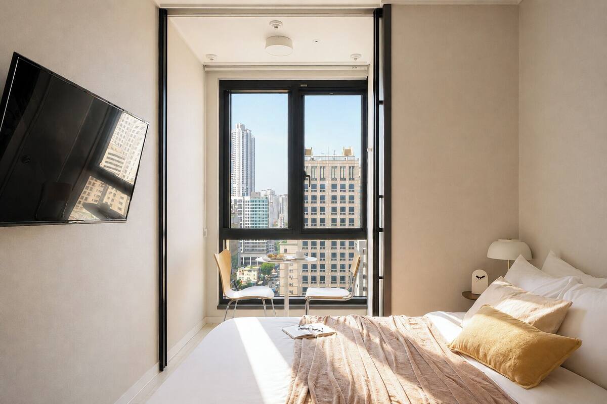 A bedroom with a bed, a TV mounted on the wall, a window with a view of buildings, and a chair by the window.