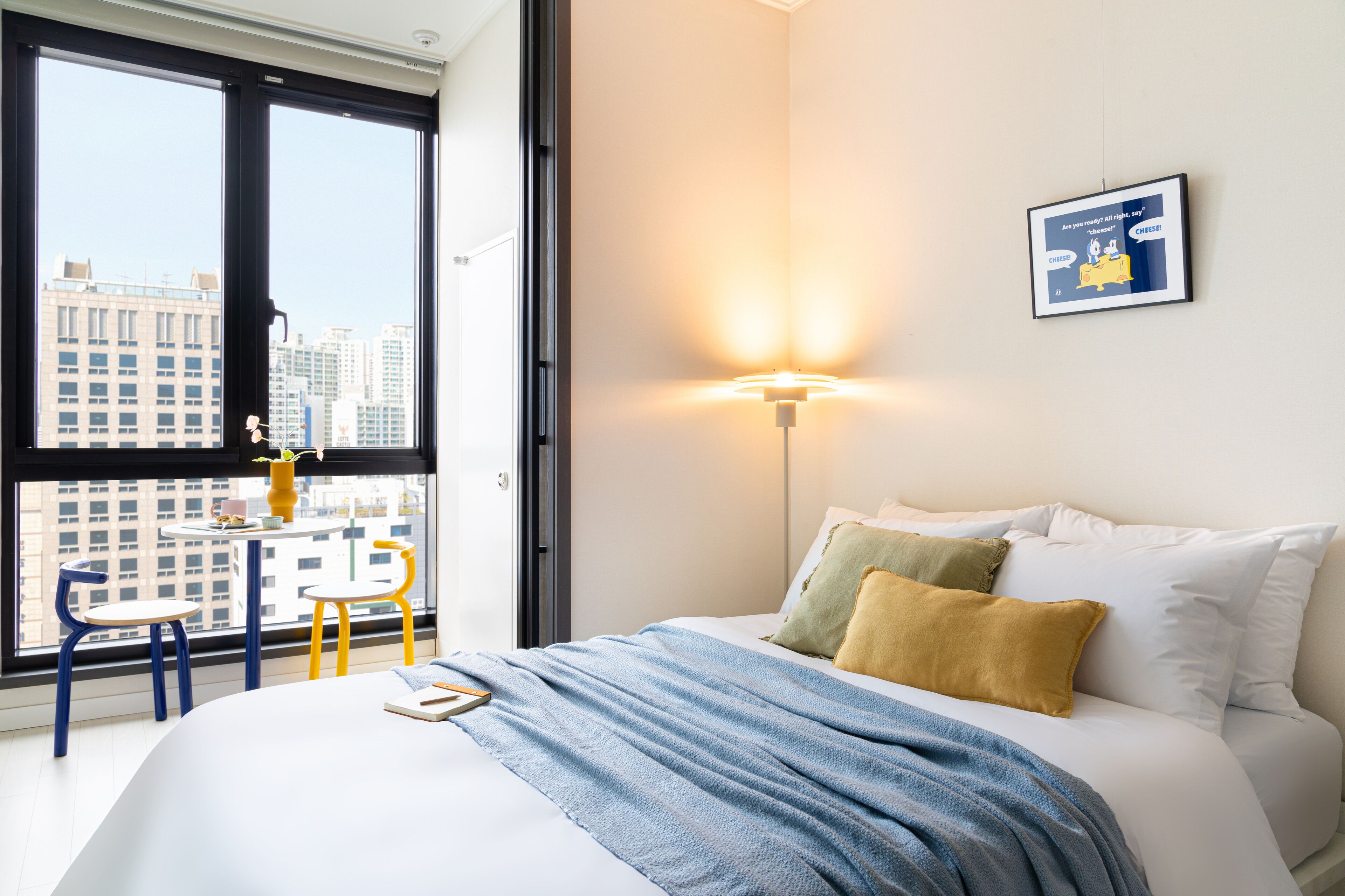 A hotel room with a large window, a bed with white and yellow pillows, a blue blanket, and a small table with chairs.