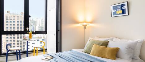 Una habitación de hotel con un ventanal grande, una cama con almohadas blancas y amarillas, una colcha azul y una pequeña mesa con sillas.