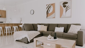 A modern living room with a sofa, coffee table, and bar stools. There is a kitchen area with a sink and a clock on the wall. - Quiet relaxing Location,2 Bedroom 2 Bathroom Guesthouse.Walking distance to CBD. (Mudgee)