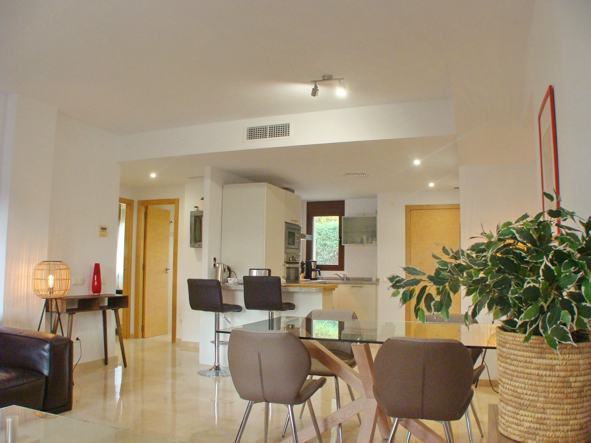 A modern living room with a glass coffee table, two armchairs, a sofa, and a dining area with a small kitchen in the background.