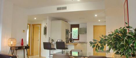 A modern living room with a glass coffee table, two armchairs, a sofa, and a dining area with a small kitchen in the background.