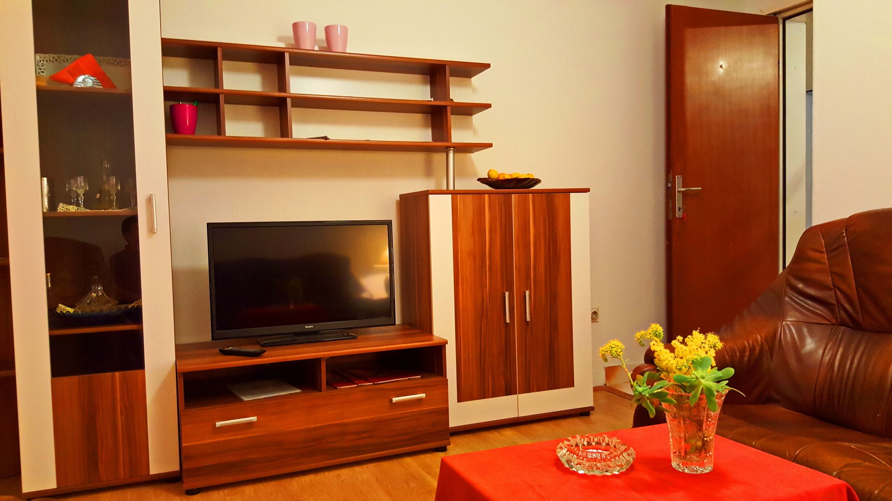 A living room with a TV, wooden entertainment unit, a red table with a vase, and a brown leather sofa.
