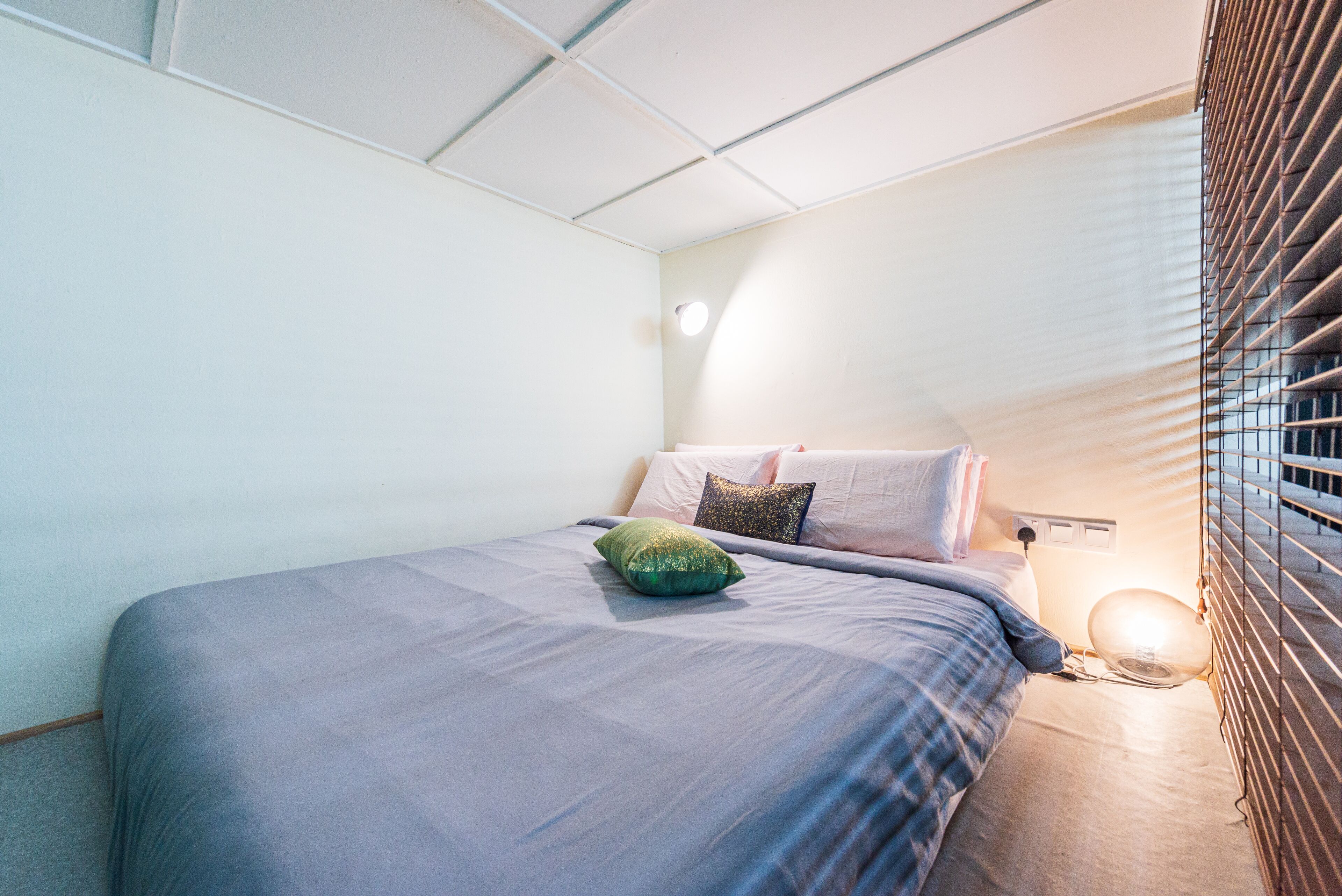 A neatly made bed with a blue bedspread and pillows, a bedside lamp, and a window with blinds.
