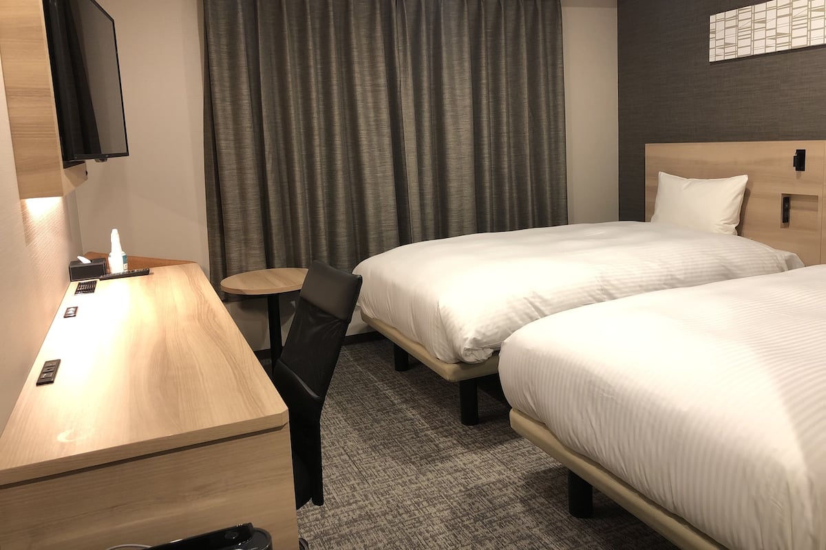 Une chambre d’hôtel avec deux lits, un bureau avec une chaise, une petite table, un téléviseur fixé au mur et une fenêtre avec des rideaux.