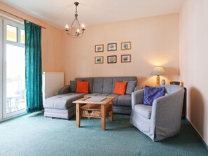A living room with a grey sofa, a wooden coffee table, and a grey armchair. There are six framed pictures on the wall, a chandelier, and a lamp on a side table. - Minervapark Whg. Mi13 (Ostseebad Boltenhagen)