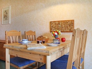 A wooden dining table with blue cushioned chairs, a woven wall art piece, and a vase with flowers and fruits.