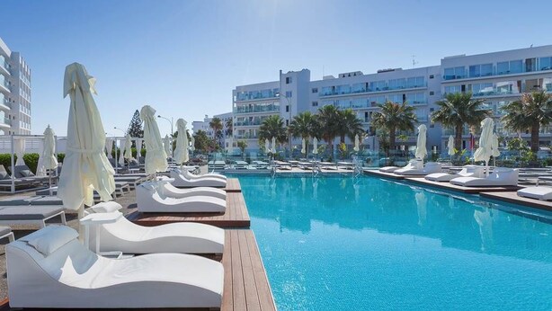 A poolside area with lounge chairs and umbrellas, a clear blue pool, and modern buildings in the background. - The Blue Ivy Hotel and Suites (Paralimni)