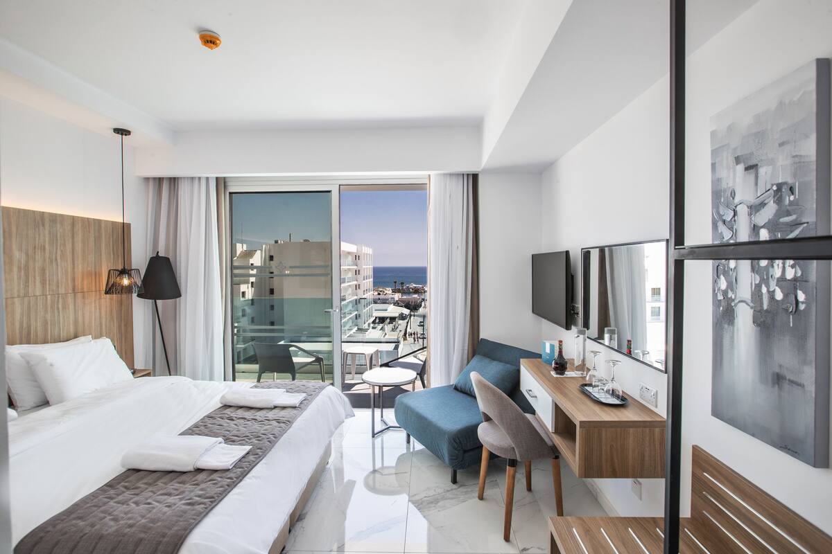 A modern hotel room with a bed, a desk, a chair, a TV, and a balcony with a view of the city.