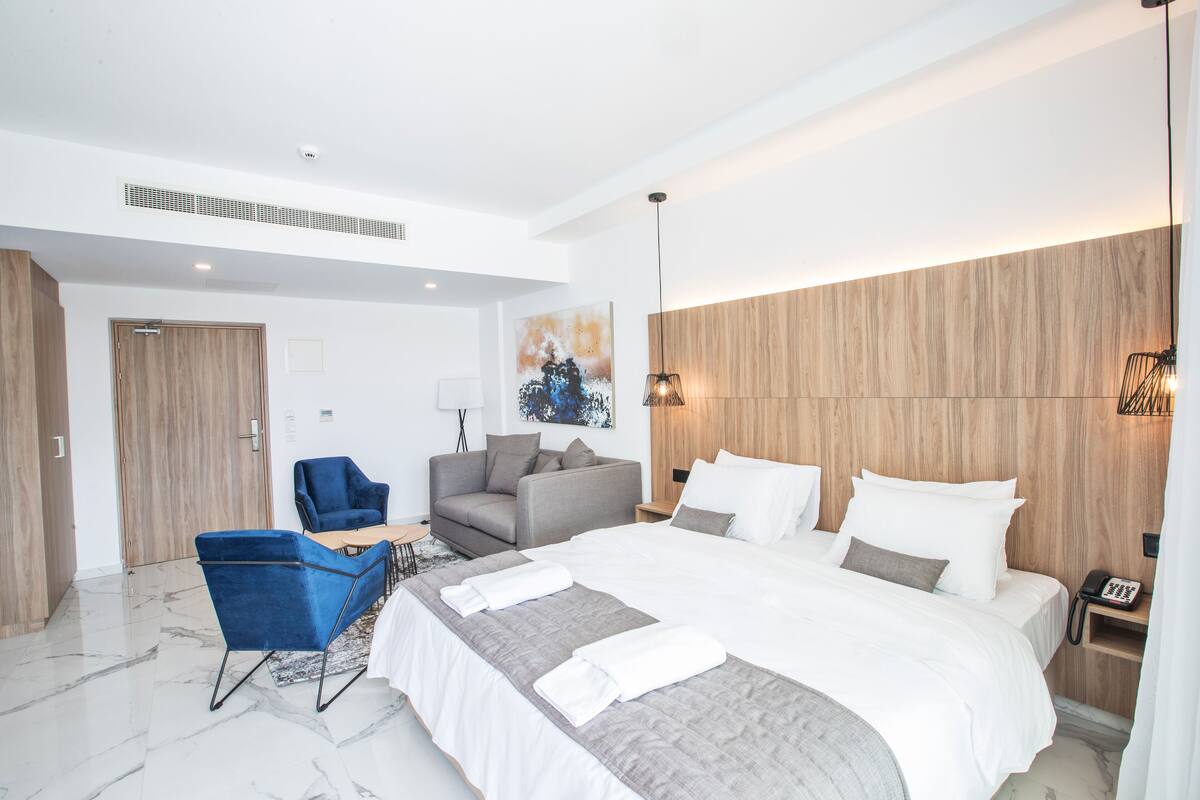 A modern hotel room with a large bed, a sofa, a blue armchair, and a wooden accent wall.