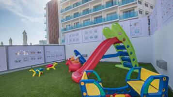 A children's play area with a slide and climbing structures, surrounded by artificial grass, against a building with balconies and decorative panels.