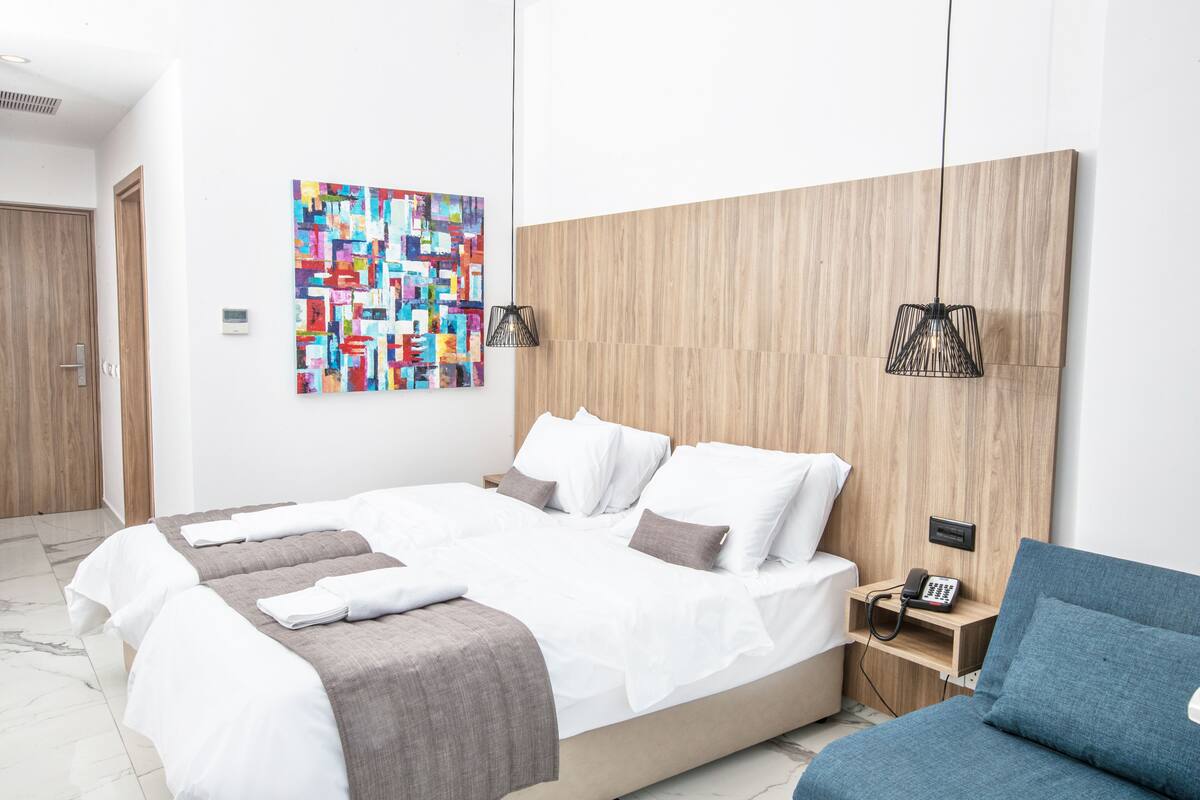A modern hotel room with a large bed, a blue armchair, a wooden headboard, a colorful abstract painting, and two pendant lights.