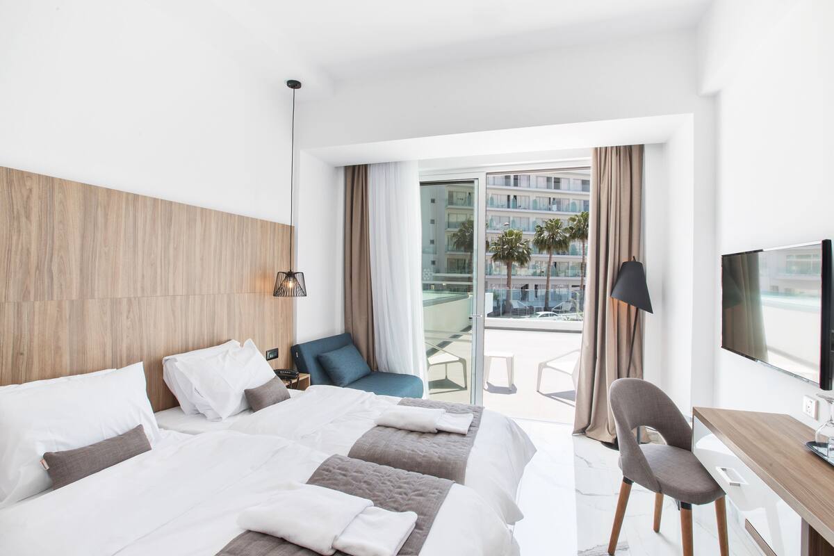 A modern hotel room with a large bed, a desk, and a balcony with a view of palm trees and buildings.