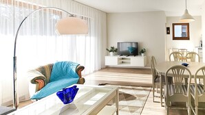 A living room with a sofa, coffee table, TV, dining table, chairs, and a large window with curtains.