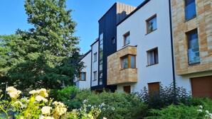 A modern multi-story residential building with a mix of white and dark siding, large windows, and a well-maintained garden with flowering plants.