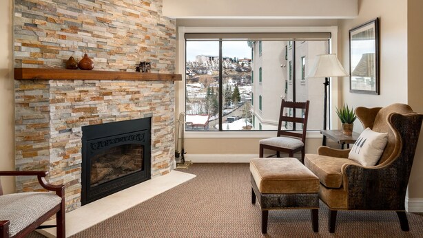 Una sala de estar con chimenea de piedra, un ventanal con vistas a edificios, una silla de madera, un banco acolchado y una mesa con lámpara.