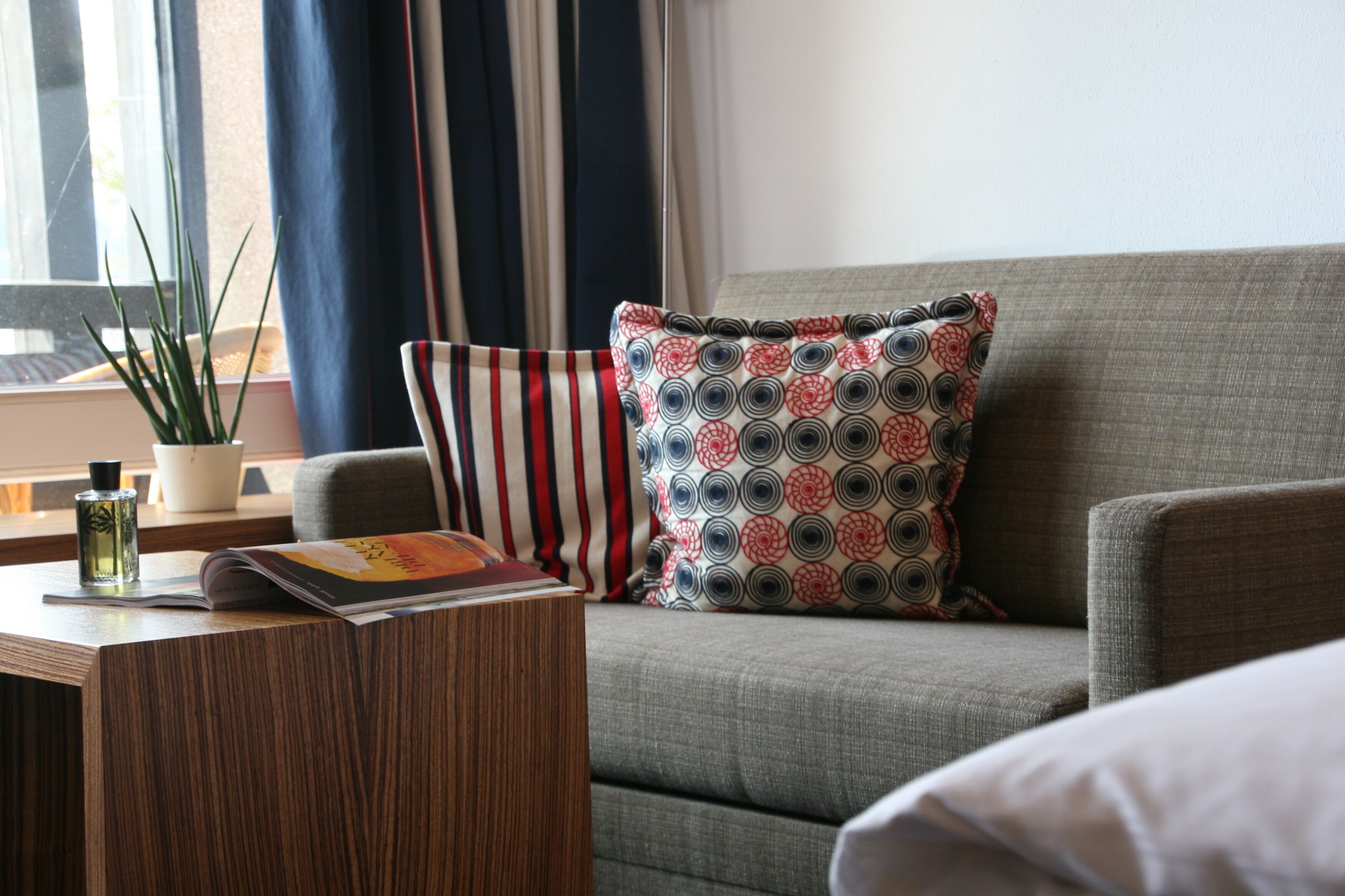 Uma sala de estar moderna com um sofá cinzento, almofadas estampadas, uma mesinha de madeira com uma revista e uma garrafa, e uma planta em vaso.
