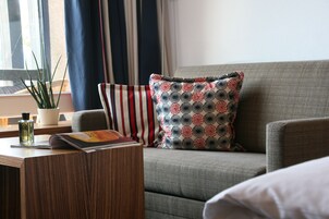 Uma sala de estar moderna com um sofá cinzento, almofadas estampadas, uma mesinha de madeira com uma revista e uma garrafa, e uma planta em vaso.