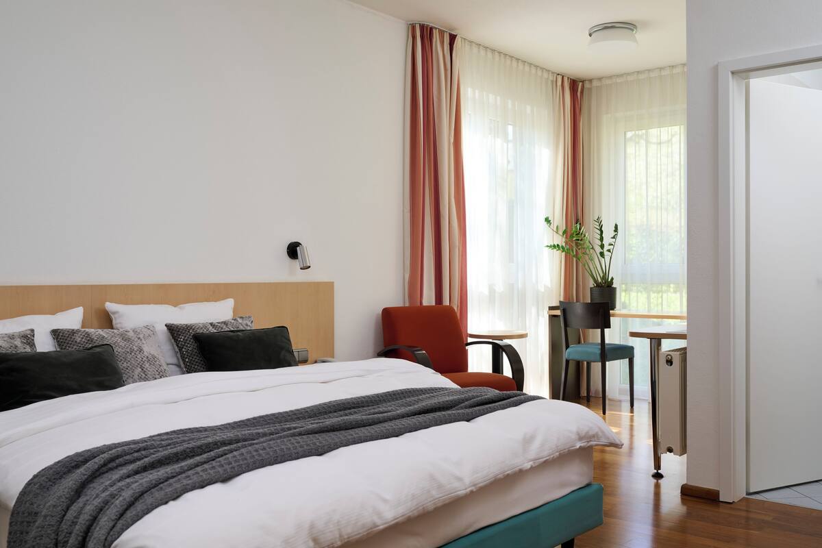 A hotel room with a large bed, a red chair, a small table with a potted plant, and a view of the outdoors through sheer curtains.