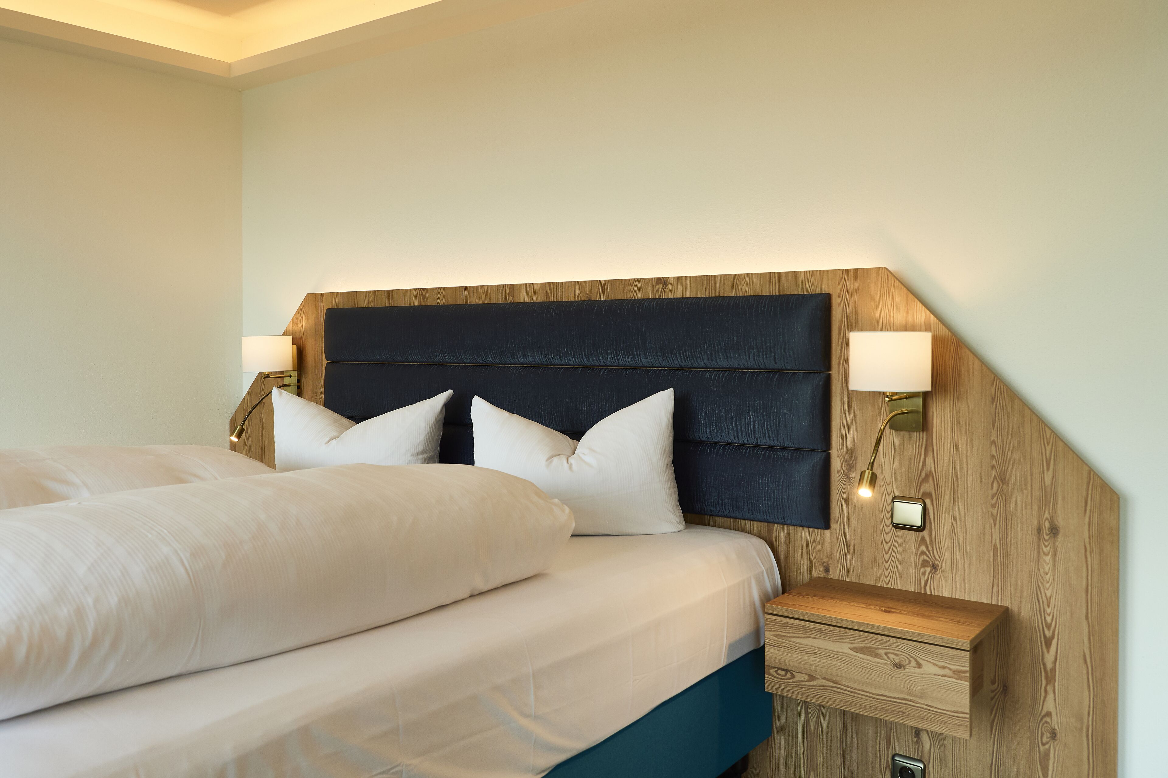 A neatly made bed with white linens and a dark headboard, flanked by two bedside lamps and a wooden nightstand with a drawer.