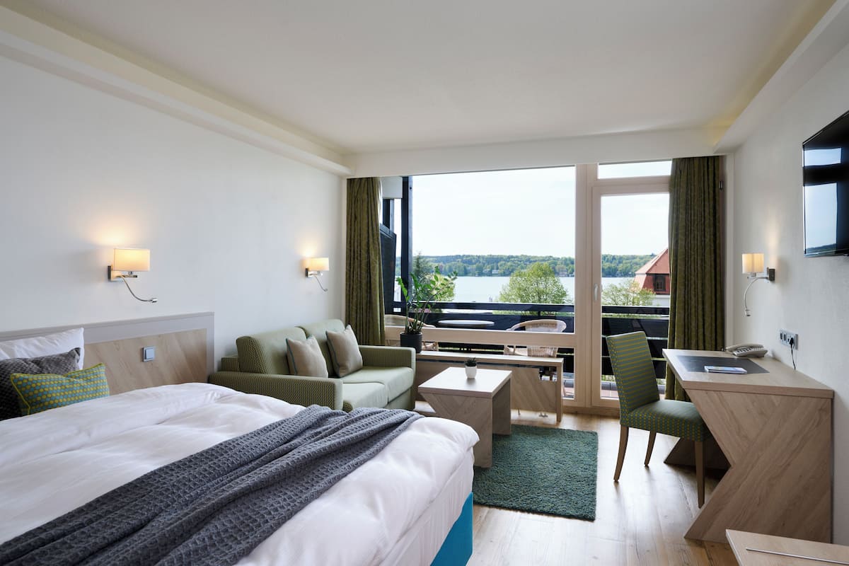 A hotel room with a bed, a desk, a chair, a sofa, and a view of a lake.