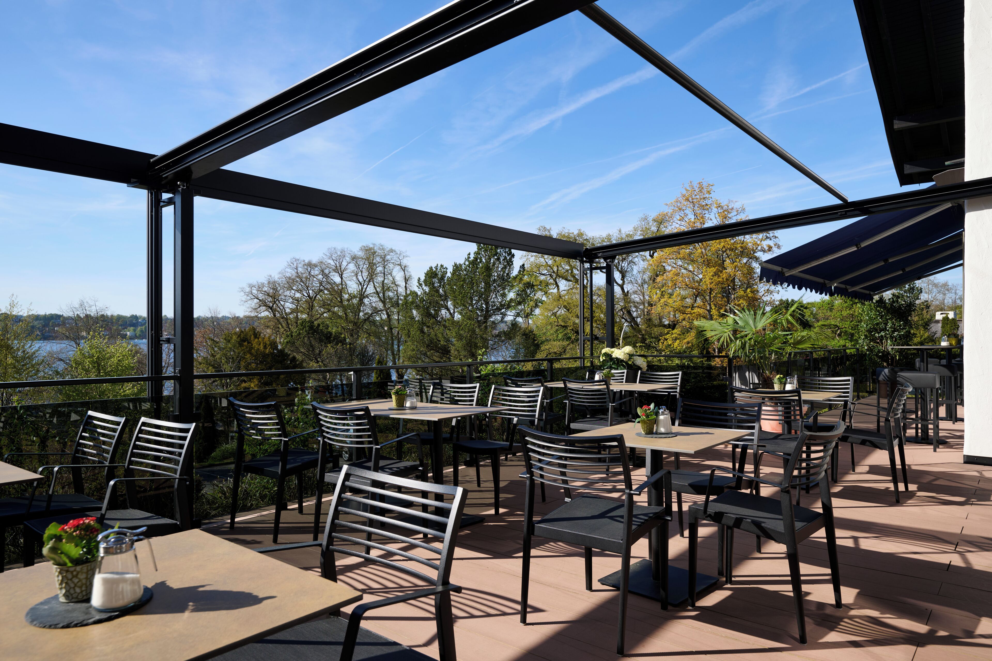 Patio esterno con tavoli e sedie, vista su alberi e un corpo d'acqua, e un'area coperta con tetto.