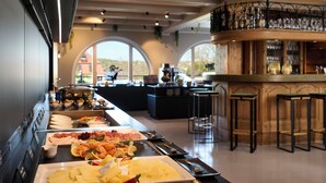 A buffet with various food items, a bar area with stools, and a view of the outside through large windows. - Hotel Schloss Berg (Berg)