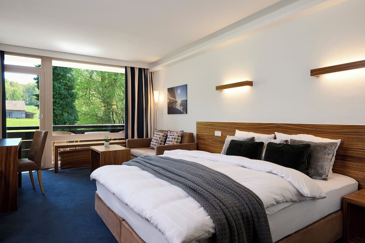 A modern hotel room with a large bed, a desk, and a chair. There is a window with a view of greenery outside.