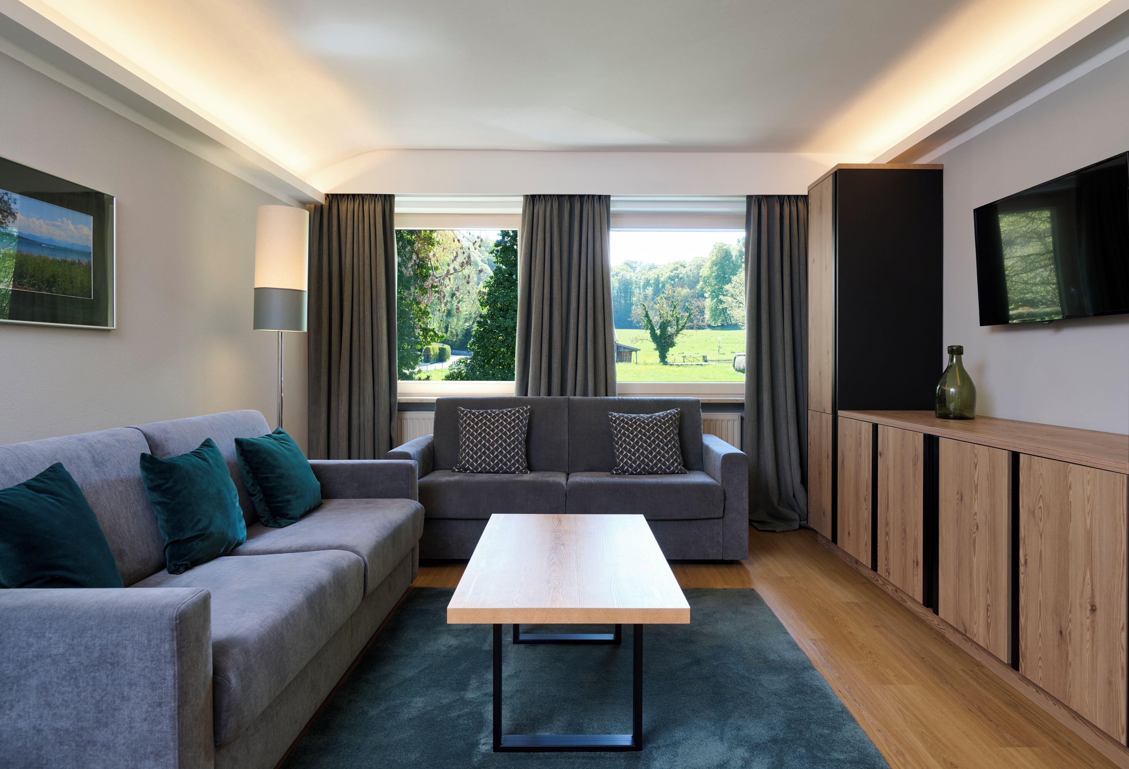 Un salon moderne avec un canapé gris, une table basse en bois, un téléviseur fixé au mur et une vue sur un paysage verdoyant grâce à de grandes fenêtres.