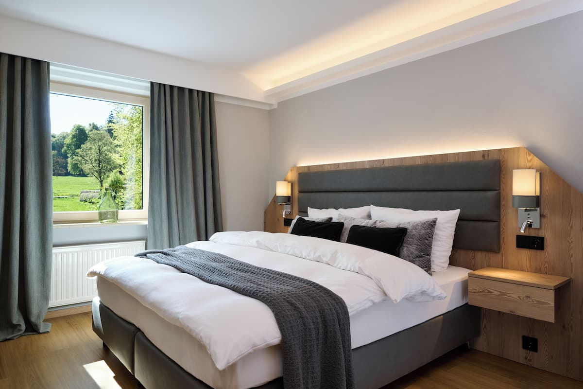 A modern bedroom with a large bed, bedside lamps, a nightstand, and a view of a green landscape through a window.