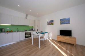 Een moderne keuken met witte kastjes, een eettafel met stoelen, een televisie op een houten kast en twee landschapsschilderijen aan de muur.