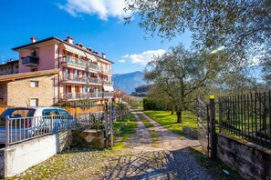 A multi-story building with balconies, a paved pathway, and a gate leading to a grassy area with trees. - Residenza Giada (Riva del Garda)