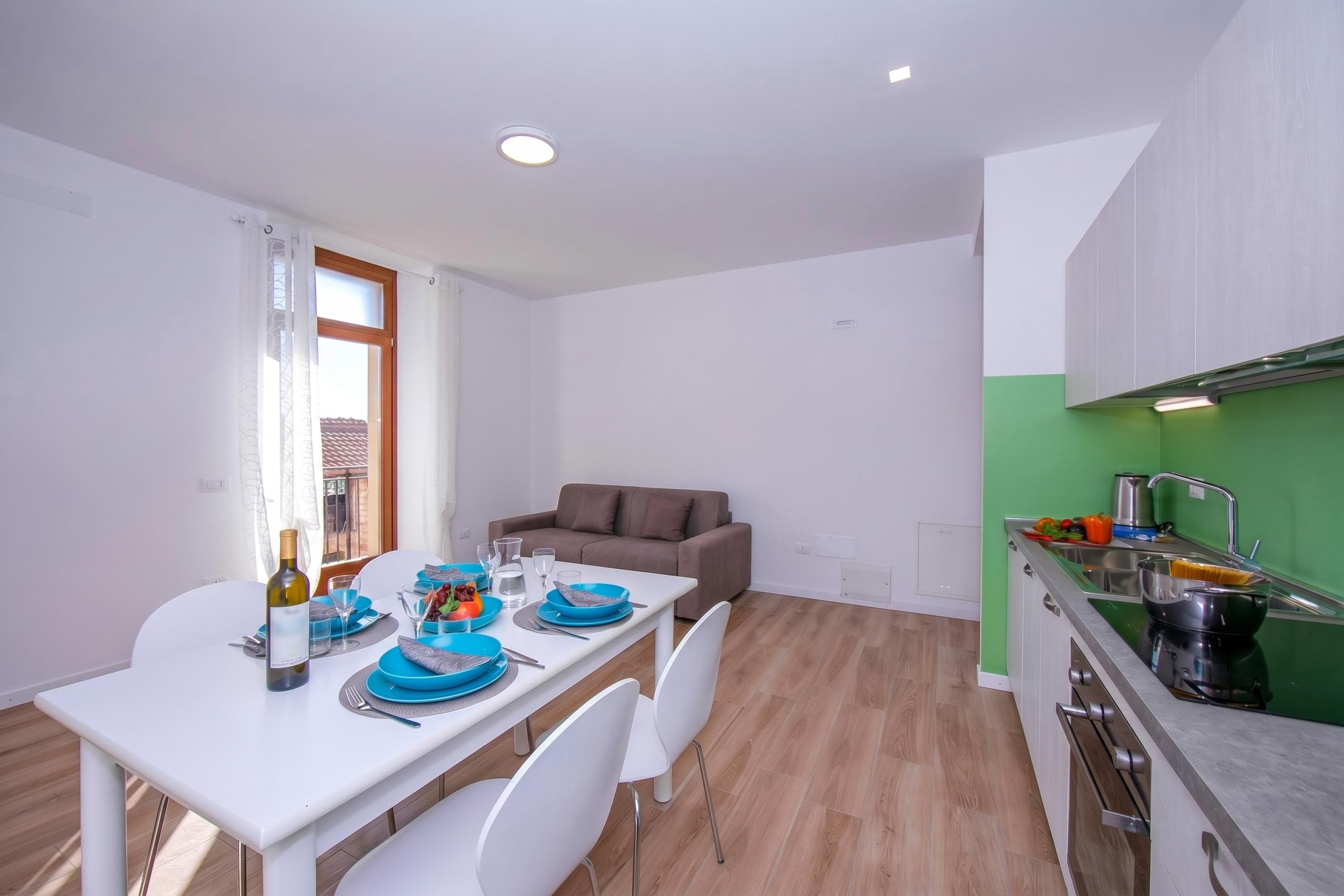 A modern kitchen with a green wall, a dining area with a white table and blue plates, and a brown sofa in the background.