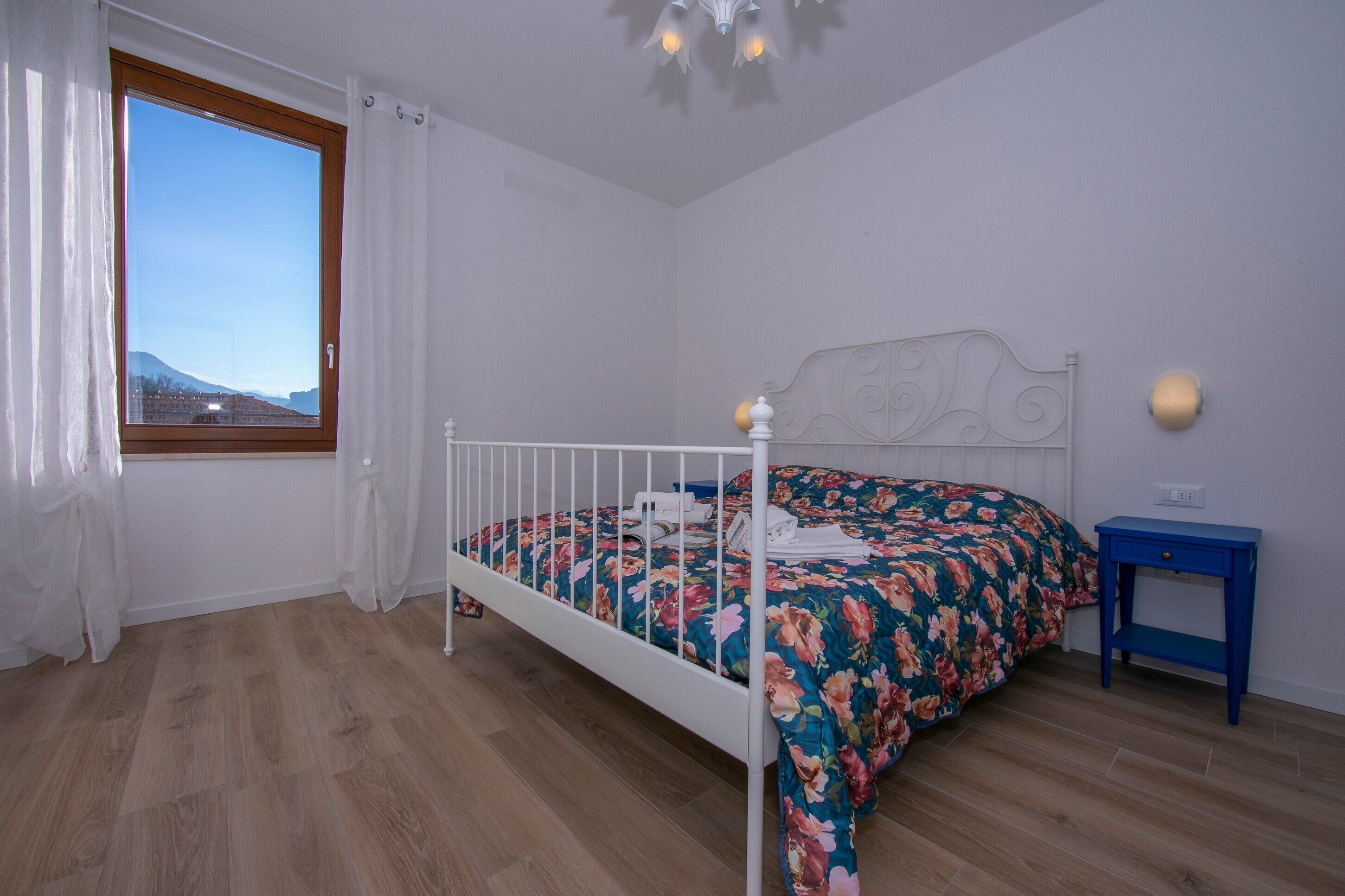 Um quarto com piso de madeira, cama com colcha florida, cabeceira branca, janela com cortinas e mesa de cabeceira azul.