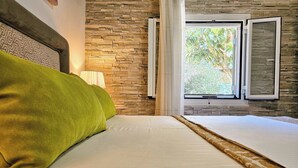 A bedroom with a stone wall, a bed with a green pillow, and a window with open shutters. - Serenity Villa Retreat in Corfu Close to Resort (Corfu)