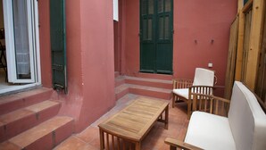 En lille patio med et træbord og to hvide stole, en rød væg og et vindue med grønne skodder.