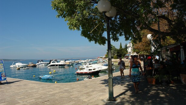 A marina with boats docked, a paved walkway, and a street with shops and people. - Apartments Villa Katarina (Selce)