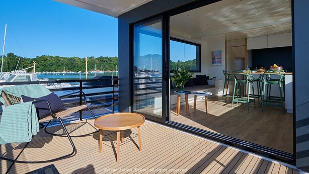 Ein moderner Balkon mit Holzboden, einer Sitzecke mit Stühlen und Tisch, und Blick auf Boote und Grünflächen.