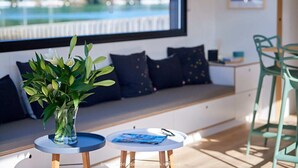 Ein modernes Zimmer mit großem Fenster und Blick auf eine Marina, ein runder Tisch mit einer Vase mit Grünpflanzen und eine Bank mit dunklen Kissen.