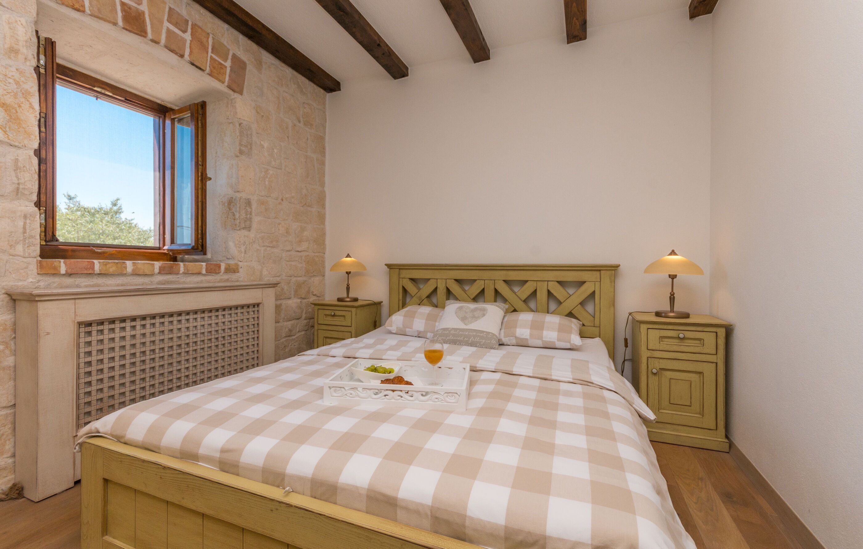 A bedroom with a checkered bedspread, a window with wooden shutters, a nightstand with a lamp, and a headboard with a heart-shaped pillow.
