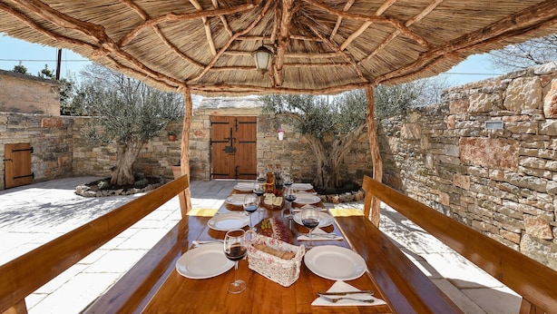 An outdoor dining area with a rustic stone wall, wooden table, and thatched roof. - Villa Calandra (Vrana)