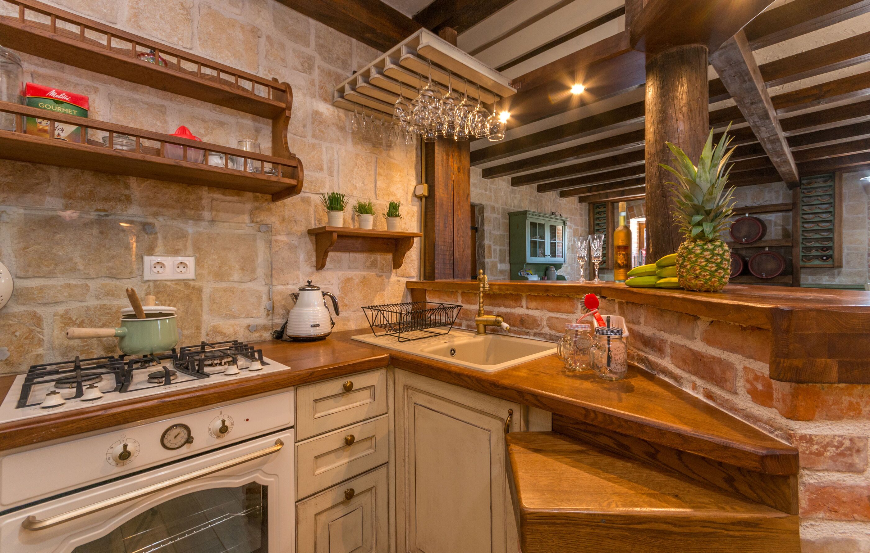 A rustic kitchen with a stone wall, wooden shelves, a gas stove, and a wooden countertop.