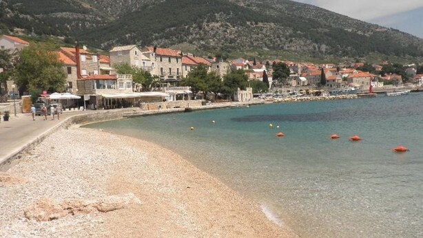 A pebble beach with clear water, buildings, and a mountain in the background. - Apartments Cukarin (Bol)