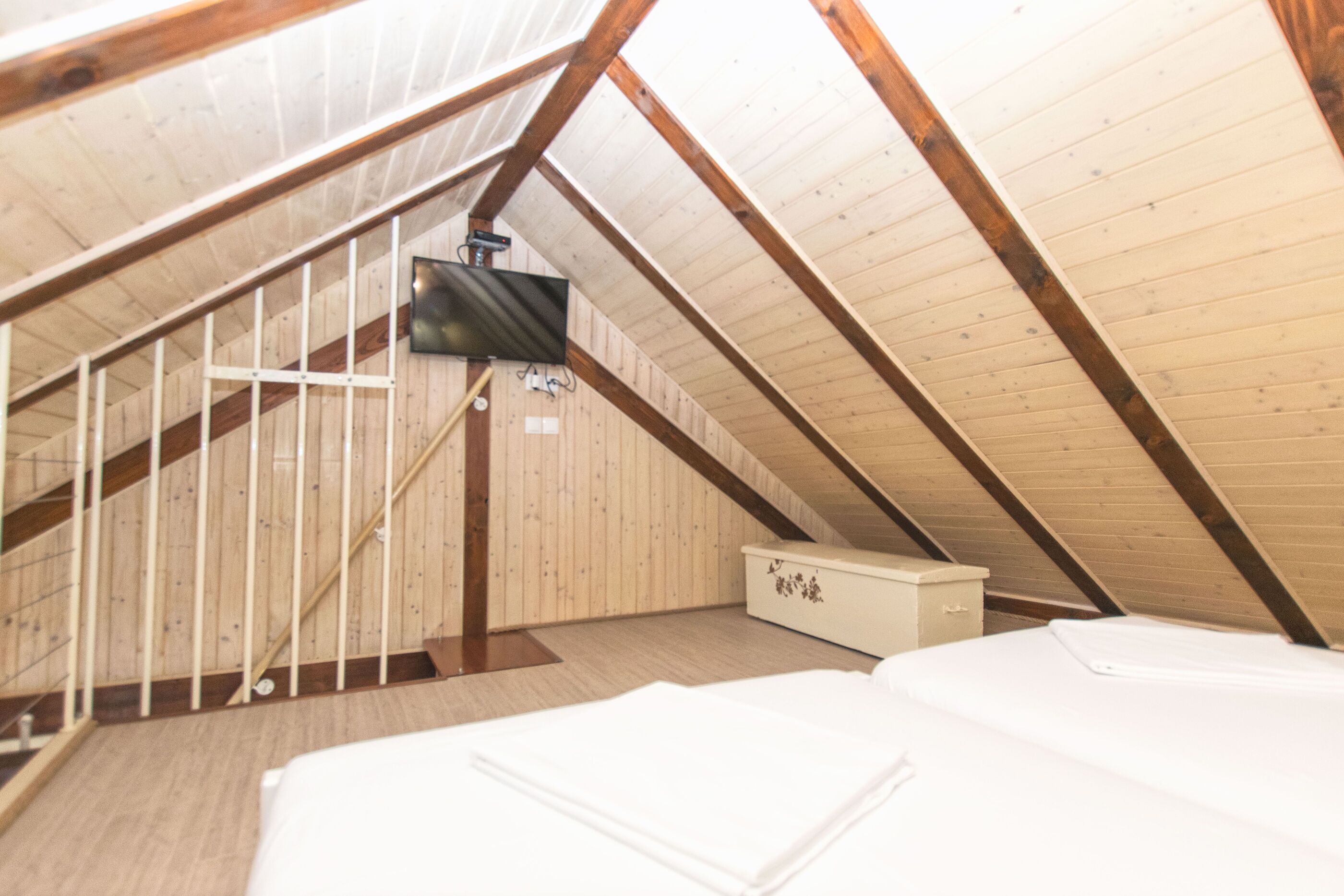 A small attic bedroom with wooden beams, a television mounted on the wall, and a white bed.