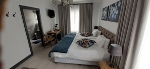 A hotel room with a bed, a desk, a chair, a TV, and a window with curtains. - Guesthouse Indongo (Swakopmund)