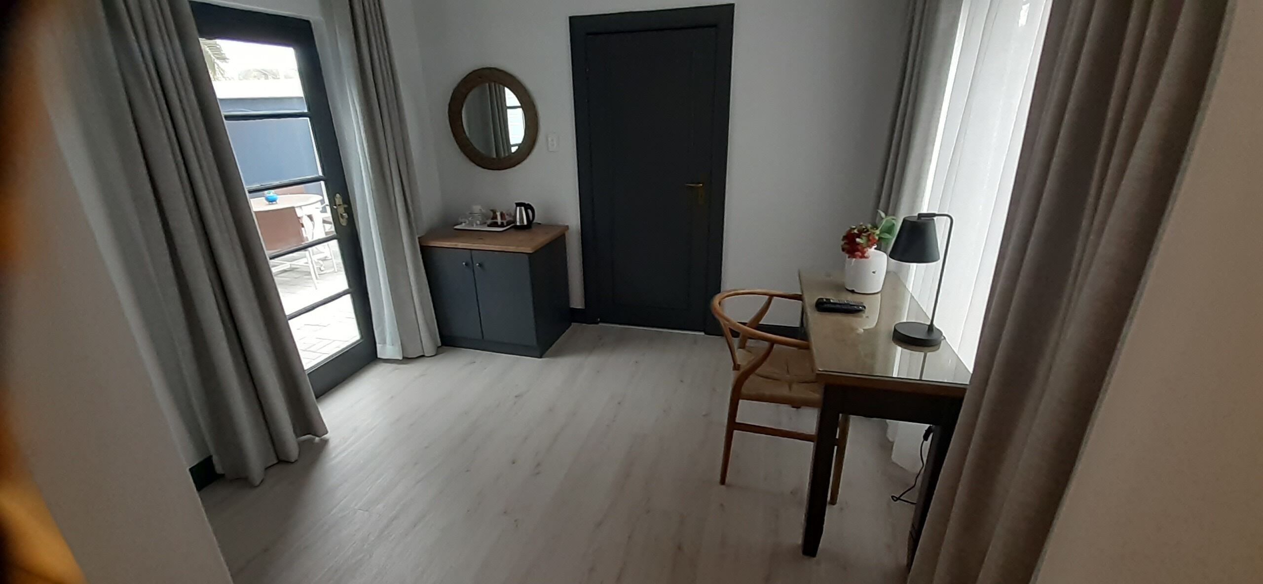 A hotel room with a wooden floor, a desk with a chair, a small cabinet, a round mirror, a door, and a window with curtains.