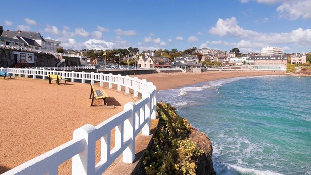 A coastal promenade with a white fence, benches, and a sandy beach.