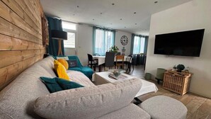 A living room with a sofa, ottoman, dining area, and a TV mounted on the wall.